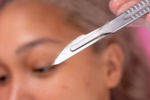 A closeup of the blade used for dermaplaning facial hair removal with client in background